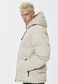 Beige Daunenjacke mit Kapuze, mit gesteppte Abschnitten, einem Reißverschluss und Kordelzug-Elementen. Mit seitlichen Taschen mit Reißverschluss.