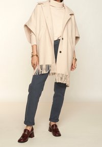 Poncho beige con flecos y dos botones, llevado sobre un cuello de tortuga claro. Combinado con pantalones azul oscuro y mocasines de tacón burdeos.