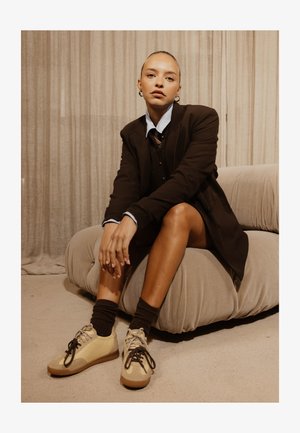 Femme aux cheveux plaqués en arrière, assise sur un canapé bas beige, portant un blazer noir, des hauts superposés, des chaussettes foncées et des baskets beiges.