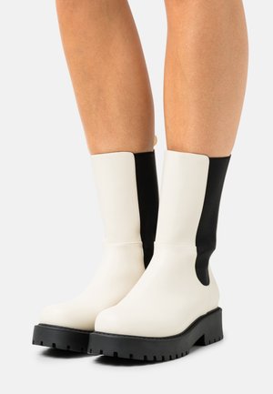 Stivaletti robusti alla caviglia con tomaia in pelle bianca e pannelli laterali elastici neri, con suola spessa in gomma nera e dettagli cuciti.