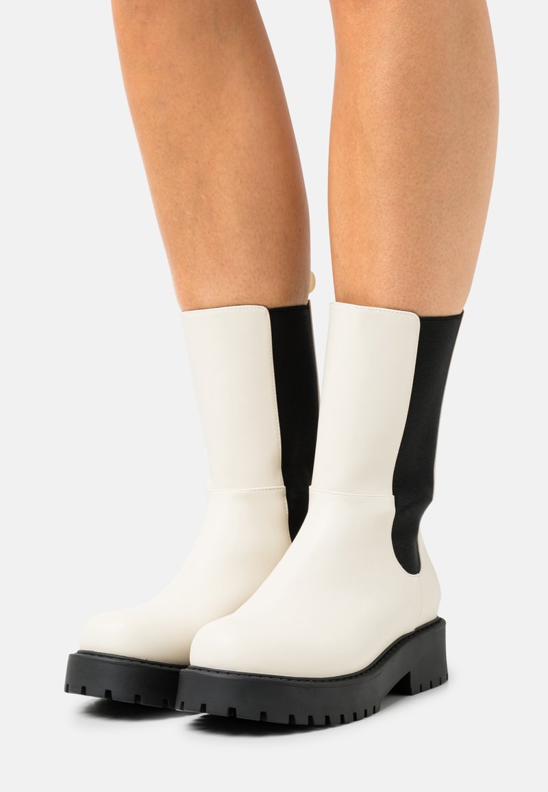 Bottines épaisses à la cheville avec une tige en cuir blanc et des panneaux latéraux élastiques noirs, dotées d'une semelle en caoutchouc noire épaisse et de détails cousus.