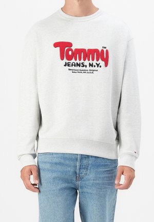 Mann trägt hellgrauen Sweatshirt mit rotem und schwarzem "Tommy Jeans, N.Y." Logo und blauen Jeans, steht vor weißem Hintergrund.