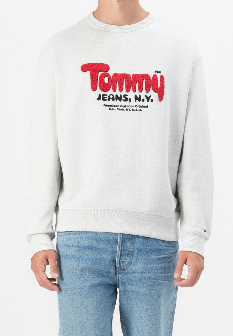 Homme portant un sweat-shirt gris clair avec le logo rouge et noir « Tommy Jeans, N.Y. » et un jean bleu, debout devant un fond blanc.