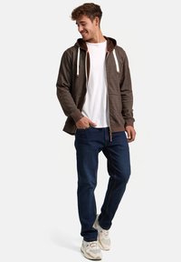Brown zip-up hoodie with a dark hood, cream drawstrings, and pocket details, paired with blue jeans and light-colored sneakers.