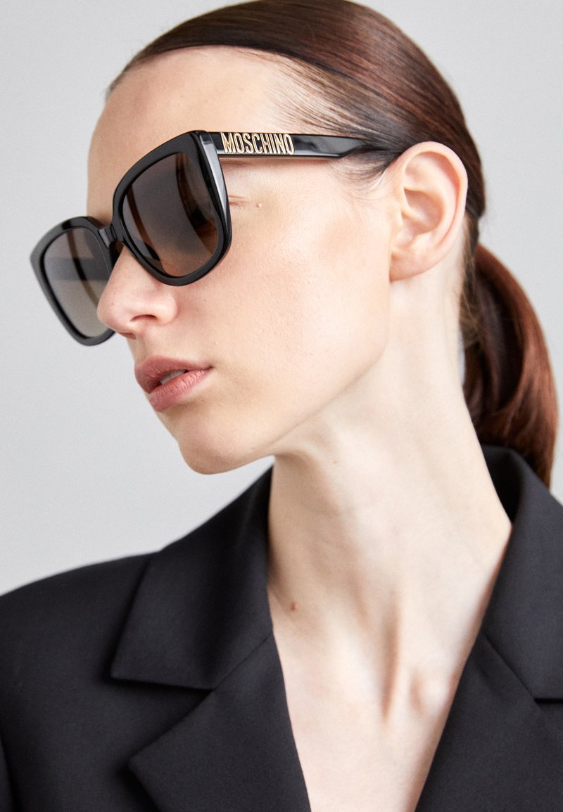 Juodos didelės saulės akiniai su tonuotais lęšiais, kuriuose yra auksinė "Moschino" logotipo detalė ant šventyklos. Lygus rėmas su kampuota forma.
