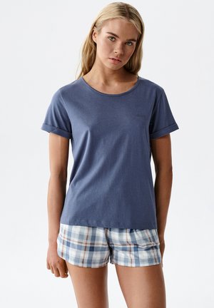 Młoda kobieta o długich blond włosach, ubrana w granatową koszulkę z krótkim rękawem oraz niebieskie, białe i brązowe szorty w kratę, stojąca na jednolitym tle.