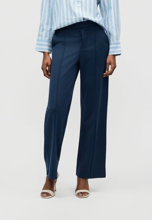 Γυναίκα που φοράει μπλε-σκούρο παντελόνι με φαρδιά μπατζάκια, ένα γαλάζιο πουκάμισο με ρίγες και μπεζ πέδιλα με ανοιχτά δάχτυλα, στέκεται μπροστά σε απλό φόντο.