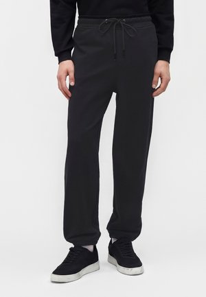 Hombre con pantalones deportivos negros con cintura de cordón y zapatillas negras con suelas blancas, de pie frente a un fondo liso.