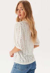 Top bianco leggero con maniche a sbuffo. Presenta motivi a occhio di pernice e una finitura testurizzata. Taglio con collo tondo e vestibilità casual.