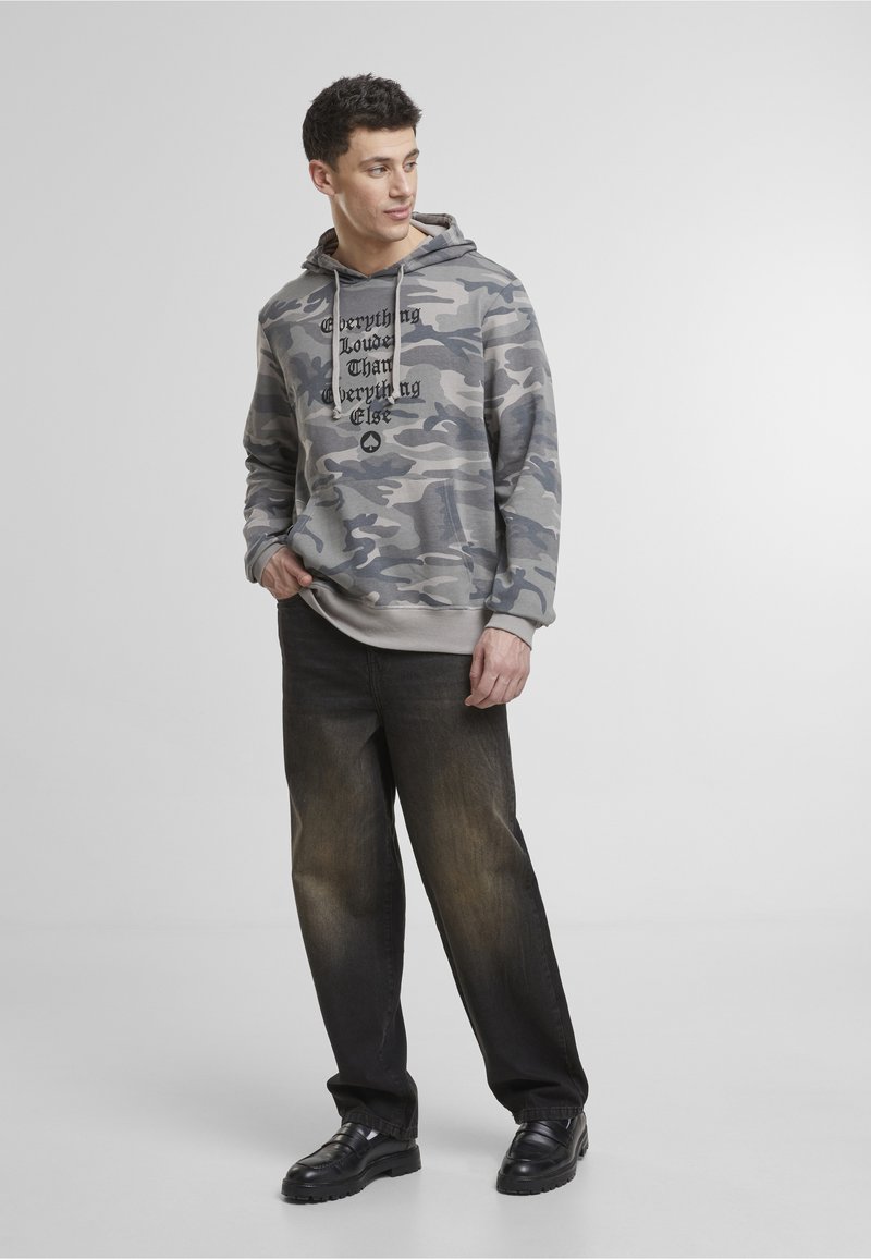 Muž oblečený v šedej kamuflážnej mikine s textom a čiernych vyblednutých džínsoch stojaci proti jednoliatemu svetlému pozadiu.