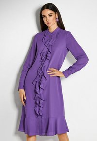 Frau mit langen schwarzen Haaren, die ein lila langärmliges Midikleid mit Rüschen an der Vorderseite trägt und vor einer weißen Wand posiert.