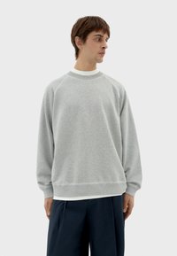 Sudadera gris claro con cuello redondo, mangas raglán largas y puños de canalé. Material suave con un corte casual y relajado. Usada sobre una camisa blanca.