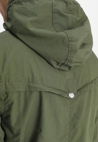 Zielona oliwkowa kurtka na zewnątrz z kapturem, wyposażona w zatrzask i widoczne przeszycia. Wykonana z lekkiego, teksturowanego materiału.