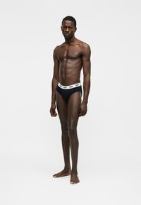 Μυώδης άνδρας σκουρόχρωμος, όρθιος, ξυπόλητος, με μαύρα εσώρουχα με λευκή ελαστική μέση με λογότυπο, σε απλό ανοιχτό φόντο.