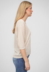 Hellbeige gestrickter Pullover mit lockerer Passform, V-Ausschnitt und kurzen Ärmeln. Hat ein strukturiertes Muster mit kleinen Löchern und wird über blauen Jeans getragen.
