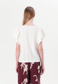 Top bianco a maniche corte con maniche a balze, tessuto a coste testurizzato e retro dritto, abbinato a pantaloni borgogna con motivo floreale.
