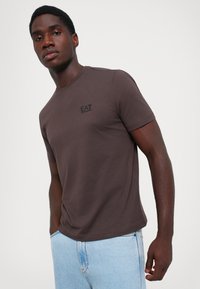 Tricou din bumbac maro, cu mâneci scurte și gât rotund, având un mic logo negru pe pieptul stâng. Purtați cu blugi de culoare albastră deschisă.