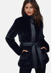 Melnas mākslīgās kažokādas mētelis ar gludu ādas priekšējo paneli, jostotu vidukli un divām sānu kabatām. Dizainam ir augsta apkakle un maiga tekstūra.