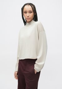 Beżowy, krótki sweter z długim rękawem, z półgolfem i prążkowaną fakturą, w połączeniu z ciemnymi bordowymi spodniami z bocznymi kieszeniami.