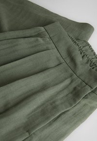 Pantaloni verde măsliniu cu talie elastică, textură vizibilă de țesătură cu model chevron și cusături adunate pe o parte.