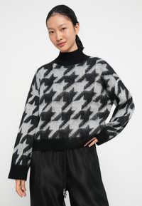 Sweter dzianinowy w czarno-szary wzór w jodełkę, z wysokim kołnierzem, długimi rękawami i luźnym krojem. Miękka tekstura, krótki fason oraz ściągacz na dole.
