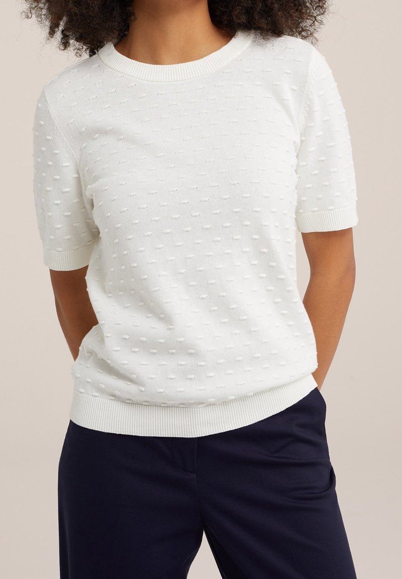 White knit sweater with textured dot patterns, round neck, and short sleeves; ribbed cuffs and hem; paired with navy pants.