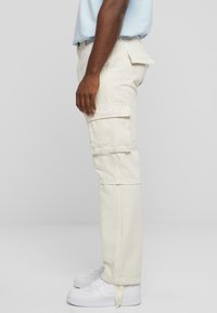 Pantalons cargo beiges avec poches latérales et coupe légèrement évasée, fabriqués en tissu robuste. Associés à des baskets blanches.