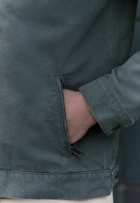 Veste en toile verte avec une poche latérale zippée, des coutures visibles et des poignets à bouton. Le tissu a une finition texturée et légèrement mate.