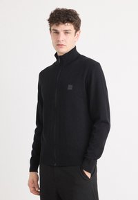 Svart zip-up tröja i ett mjukt tyg. Har en hög krage, långa ärmar och ett litet logopatch på bröstet. Idealisk för lager på lager.