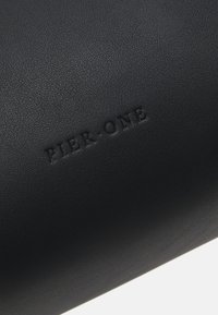 Sac cylindrique noir en synthétique fabriqué dans un matériau texturé. Présente un logo "PIER-ONE" en relief, un design minimaliste et une finition mate.