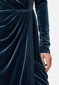 Samtkleid in tiefem Teal mit langen Ärmeln, ausgestattet mit Drapierungsdetails und kontrastierenden glatten Nähten. Die weiche Textur verstärkt die Eleganz.