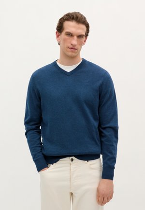 Marineblauwe v-hals trui van zacht materiaal. Heeft lange mouwen en geribbelde manchetten. Draagt over een wit t-shirt met crèmekleurige broek.