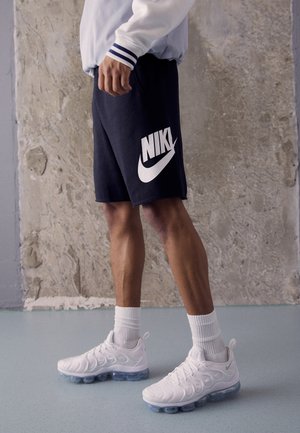 Pantalones cortos de Nike en tejido azul oscuro con un gran logo blanco, combinados con zapatillas blancas que presentan un diseño texturizado y suela aérea transparente.