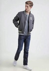 Ung mann i grå varsityjakke, grå t-skjorte, mørkeblå jeans og hvite sneakers, står med hendene i lommene og ser til venstre.