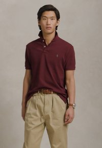 Mlad moški nosi bordo polo majico, bež nabran hlače s pletenim pasom in ura na zapestju, stoji pred enotno ozadje.