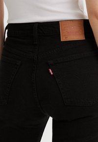 Primer plano de una persona que lleva unos vaqueros Levi's 501 negros con bolsillos traseros, una etiqueta de marca de cuero marrón y una etiqueta roja de Levi's, combinados con una camiseta blanca.