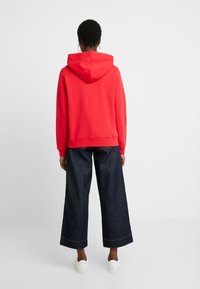 Felpa rossa con cappuccio e tasca a marsupio frontale, abbinata a pantaloni a gamba larga in denim scuro e sneakers bianche. Tessuto dalla texture liscia.