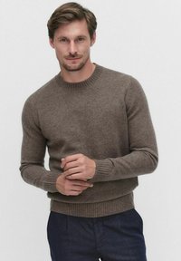 Ruskea, ribbiknitti villapaita, jossa on pyöreä kaula-aukko, pitkät hihat ja tiukka, tekstuurinen kangas. Pukeutunut tummien housujen päälle.