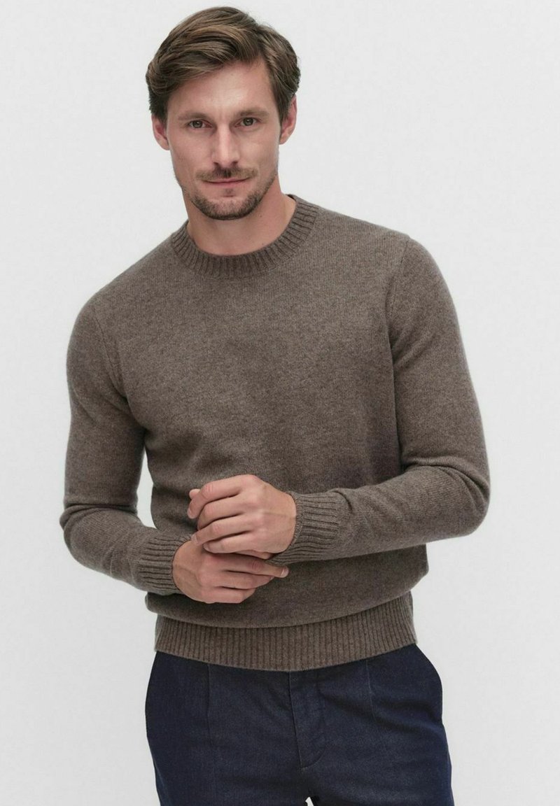 Ruskea, ribbiknitti villapaita, jossa on pyöreä kaula-aukko, pitkät hihat ja tiukka, tekstuurinen kangas. Pukeutunut tummien housujen päälle.