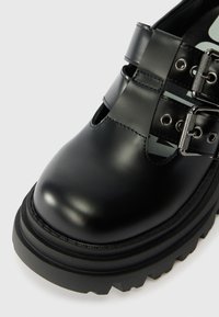Černé lesklé kožené boty s výřezy a dvojitým sponkovým detailem. Silná gumová podrážka s drsným vzorkem. Zaoblený design špičky.
