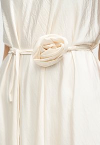 Gros plan d'une ceinture en tissu crème nouée autour de la taille avec un grand détail en tissu en forme de rose sur une robe assortie.