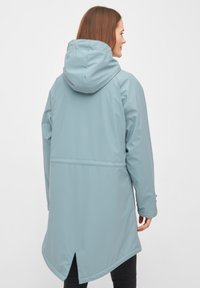 Chaqueta impermeable azul claro con capucha, que presenta una ventilación trasera y un dobladillo más largo. Está fabricada con un material sintético suave y tiene un diseño con detalles mínimos.