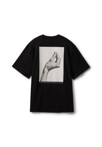 Camiseta de algodón negra de corte holgado, con una imagen impresa de una mano formando un gesto en la parte posterior. Texto sencillo debajo del gráfico.