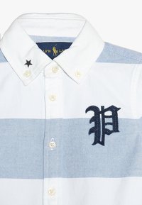 Bílá a světle modrá pruhovaná košile s tmavě modrou vyšívanou značkou "P" a černou hvězdou na límci. Zapínání na knoflíky.