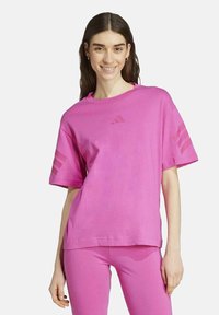 Woman smiling wearing a matching bright pink Adidas t-shirt and leggings with logo on chest and stripes on sleeves against white background.