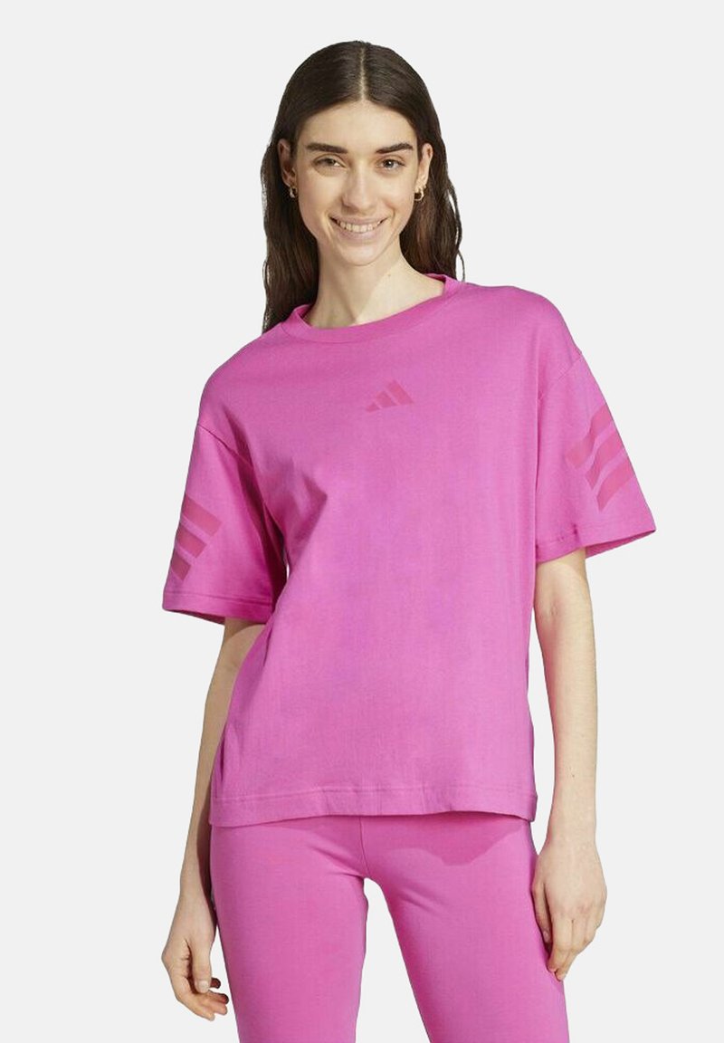 Woman smiling wearing a matching bright pink Adidas t-shirt and leggings with logo on chest and stripes on sleeves against white background.