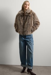 Chaqueta de piel sintética en marrón claro con una textura suave y un gran cuello, combinada con jeans azules holgados y botas de tobillo negras brillantes y puntiagudas.