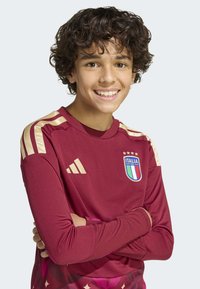 Teenage boy with curly hair smiling, wearing a maroon Italy football jersey with gold stripes and team crest, arms crossed.