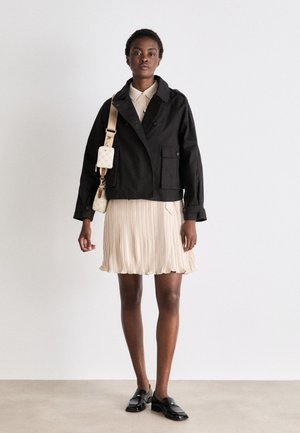 Black cropped jacket with a collar and two pockets, paired with a pleated beige dress. Black loafers and a small, light bag are also present.