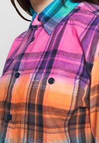 Ruudullinen flanellikangas-paita, jossa on vaaleanpunaisia, violetteja ja oransseja sävyjä; sisältää turkoosin kauluksen ja mustat napit; pehmeä rakenne.